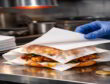 Gloved hand adds a second wrapper over a grease-soaked fried chicken sandwich on a commercial kitchen counter.