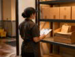 Worker checks paper bag storage on stockroom shelves beside organized cartons in a back-of-house storeroom.