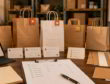 Paper bag planning desk with sample bags, supplier cards, stock triggers, and packaging swatches in a warm office.