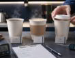 Three steaming paper cups on a cafe test station with timer, thermometer, checklist, and hands placing a lid.